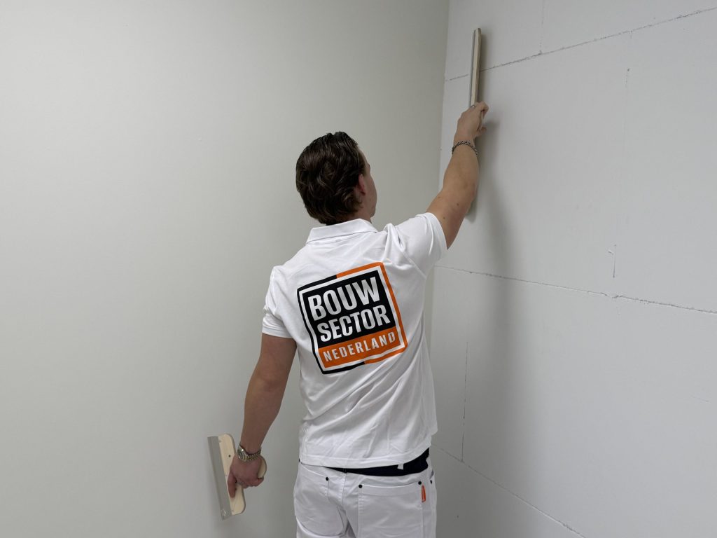 Stukadoor in wit werkshirt met logo trekt een verse pleisterlaag in de hoek van een kamer glad