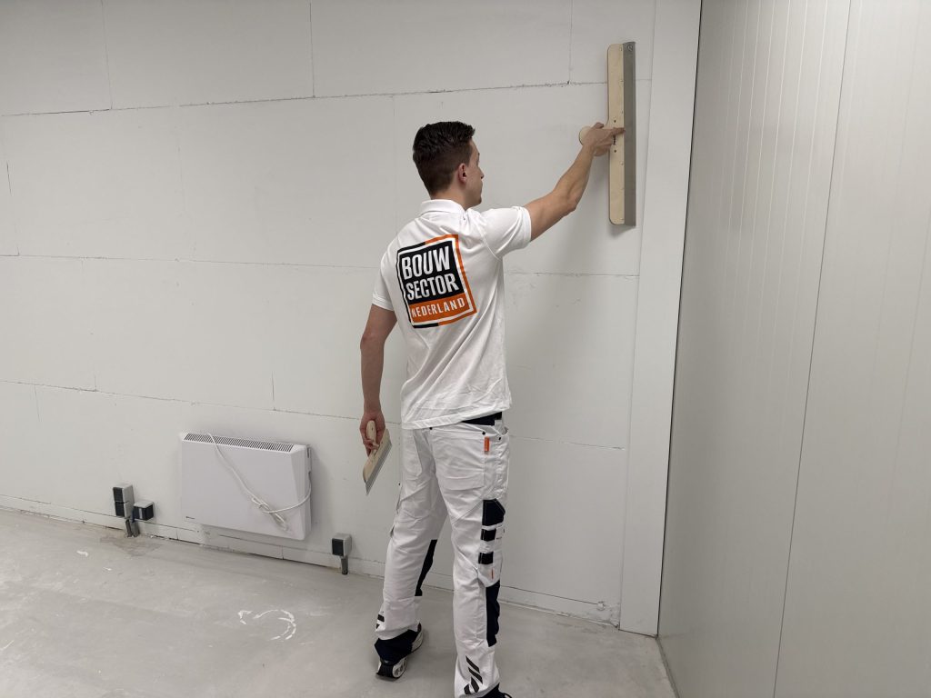 Stukadoor in witte werkkleding staat voor een kale wand en trekt de spaan omhoog langs de hoek
