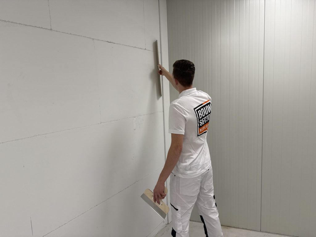 Stukadoor zet een lange spaan verticaal tegen een wand naast een paneelwand