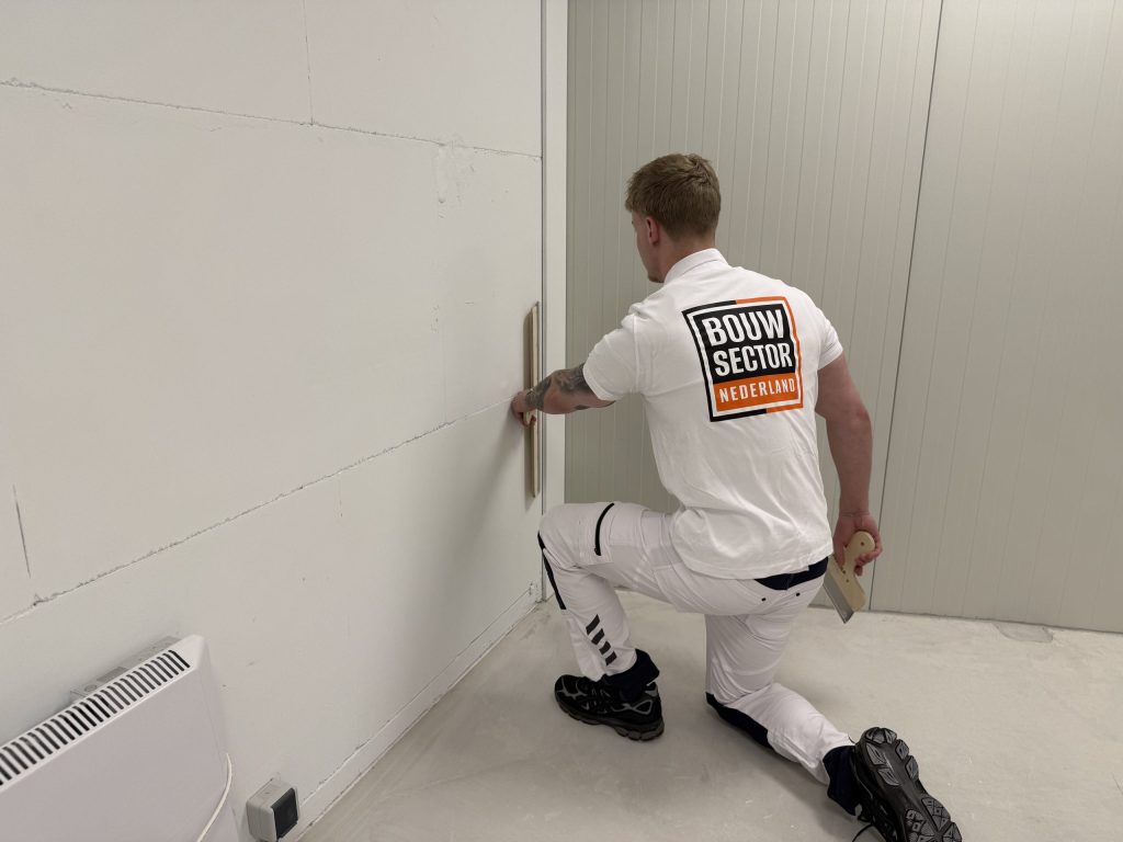 Stukadoor zit op een knie en werkt de onderkant van een lange wand strak af tot vlak bij de vloer