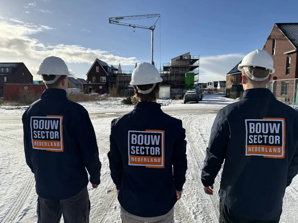 Drie medewerkers in donkere bedrijfsjassen kijken naar nieuwe bakstenen woningen in aanbouw