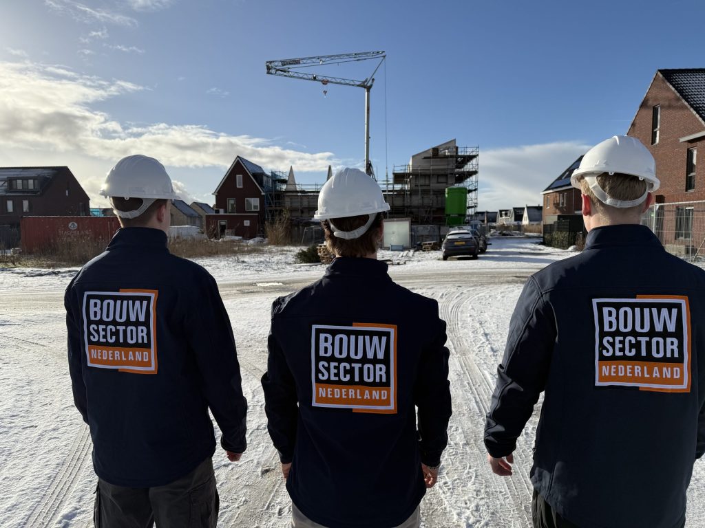 Drie vakmensen van Bouwsector Nederland met witte helm lopen een straat met nieuwe huizen en een bouwkraan in
