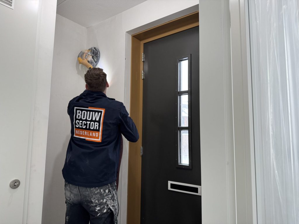 Vakman in Bouwsector-werkjas schuurt de wand boven een voordeur met een schuurmachine op een lange steel