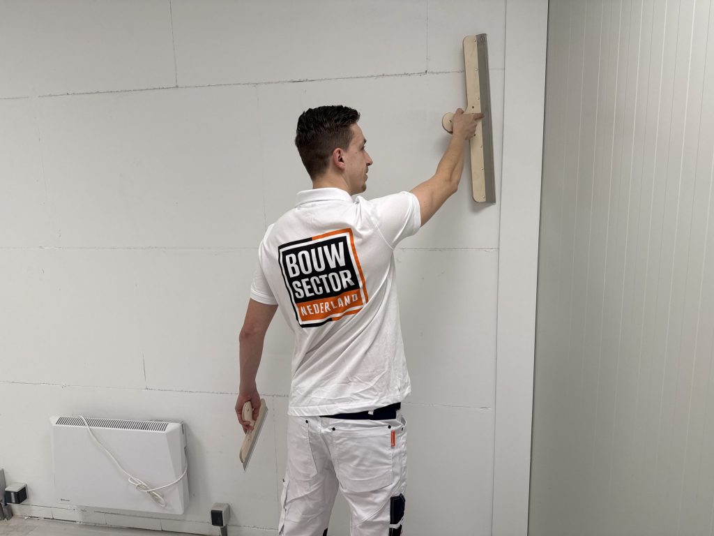 Stukadoor van Bouwsector Nederland kijkt opzij terwijl hij met een lange spaan een wand vlak trekt